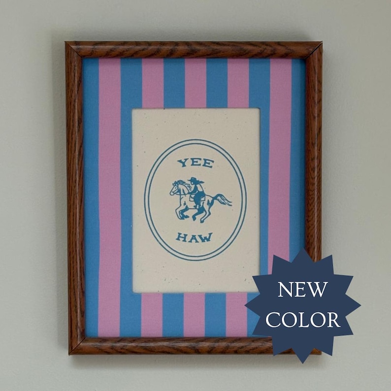 May include: A framed print featuring a cream-colored center with a blue oval border, a blue illustration of a person riding a horse, and the text YEE HAW. The background consists of vertical blue and pink stripes, all enclosed in a dark wood frame.