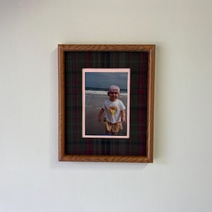 May include: A rectangular wooden picture frame featuring a dark green and red plaid fabric matting. The frame contains a photograph of a small child wearing a white shirt and a pink hat standing on a sandy beach near the ocean waves.