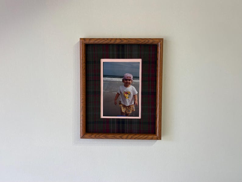 May include: A rectangular wooden picture frame featuring a dark green and red plaid fabric matting. The frame contains a photograph of a small child wearing a white shirt and a pink hat standing on a sandy beach near the ocean waves.