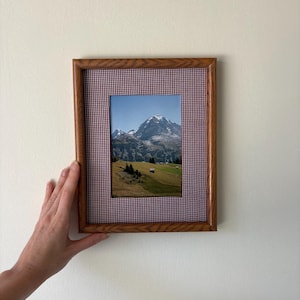 May include: A rectangular wooden picture frame featuring a fabric mat with a small red and white gingham check pattern. The frame contains a photograph of a snow-capped mountain peak rising above a green grassy landscape with small trees.