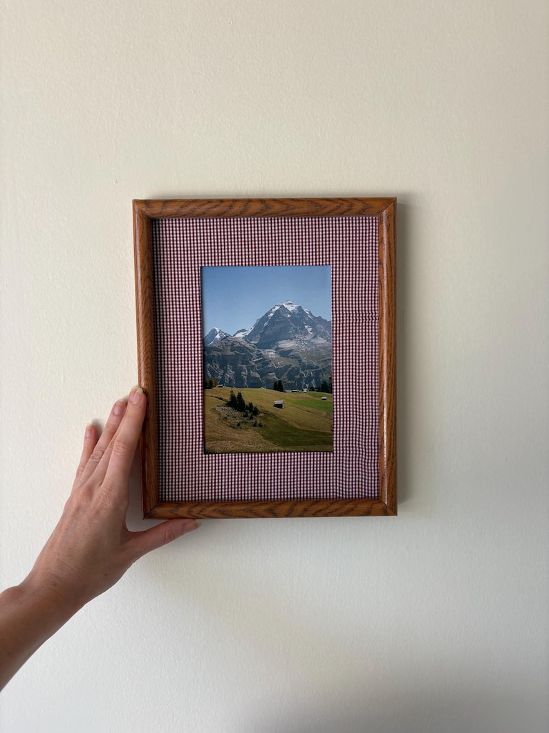 May include: A rectangular wooden picture frame featuring a fabric mat with a small red and white gingham check pattern. The frame contains a photograph of a snow-capped mountain peak rising above a green grassy landscape with small trees.