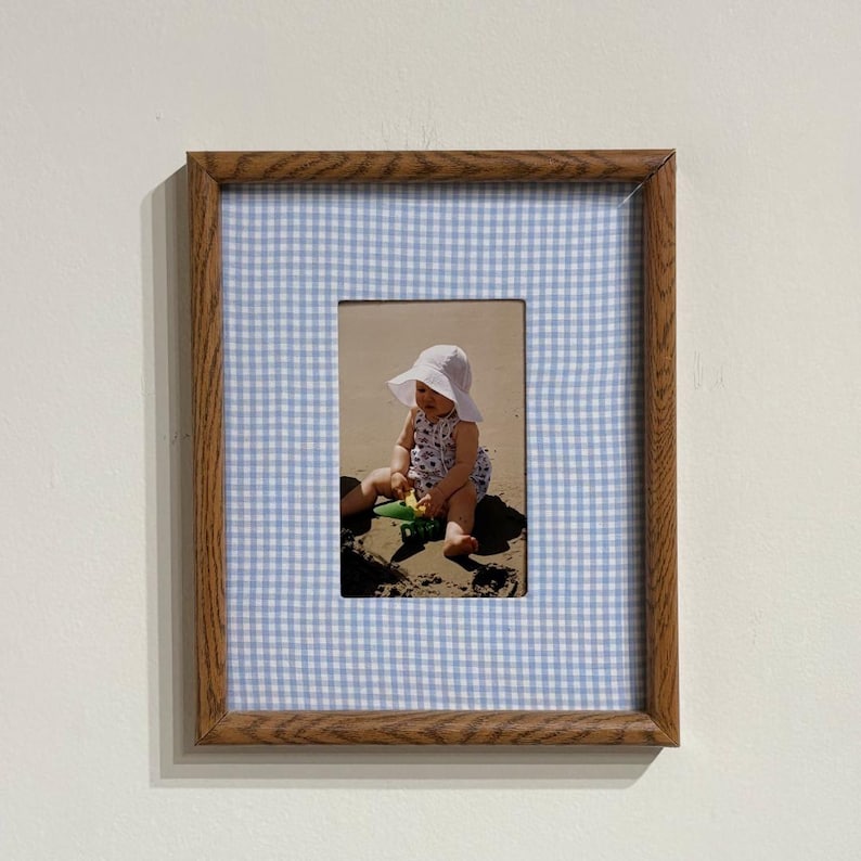 May include: A rectangular wooden picture frame featuring a light blue and white gingham fabric mat. The frame contains a photograph of a small child wearing a white sun hat and a patterned outfit, sitting on sand and playing with a green toy.