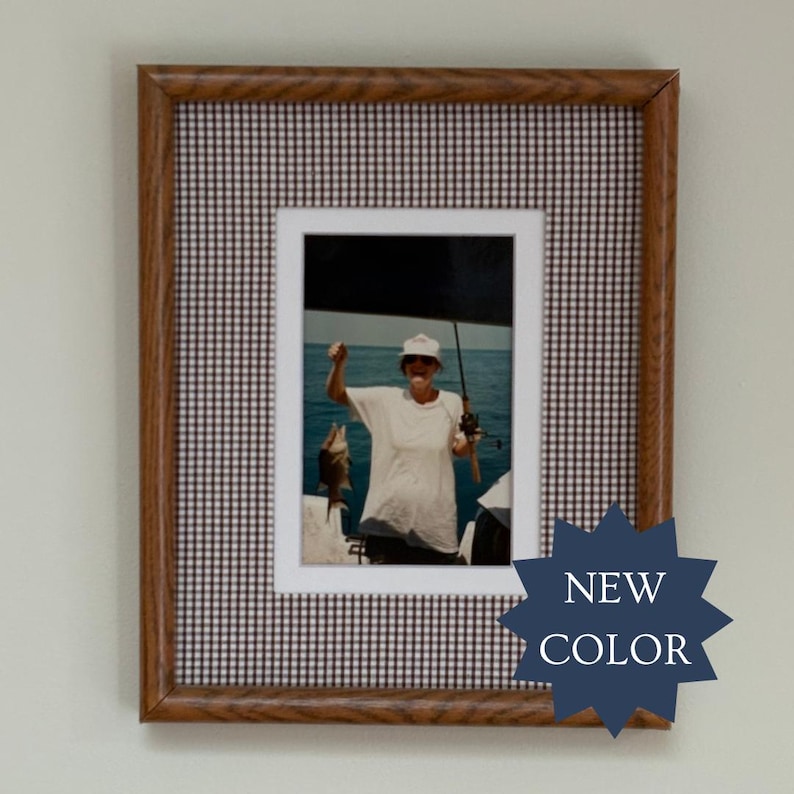 May include: A rectangular wooden picture frame featuring a brown wood grain finish and a black and white gingham matting. The frame contains a photograph of a person wearing a white shirt and hat, holding a fishing rod and a caught fish.