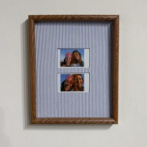May include: A rectangular wooden picture frame featuring a blue and white pinstriped mat board. The frame contains two stacked rectangular photo openings, each displaying a photograph of a person wearing a pink party hat, centered within the mat.