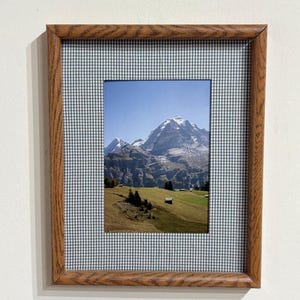 May include: A rectangular picture frame with a medium-brown wood grain finish and a gray and white gingham fabric mat. The frame contains a photograph of a snow-capped mountain peak rising above a green grassy meadow with small wooden structures.