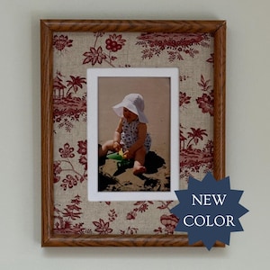 May include: A rectangular wooden picture frame featuring a beige fabric mat with a red floral toile pattern. The frame contains a photograph of a child wearing a white sun hat sitting on a sandy beach. A dark blue star-shaped badge reads NEW COLOR.