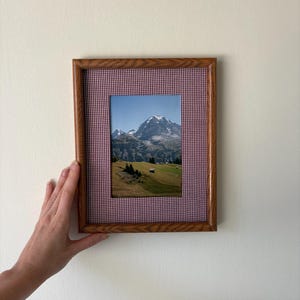 May include: A rectangular wooden picture frame featuring a red and white gingham fabric matting. The frame contains a photograph of a snow-capped mountain peak rising above a green grassy landscape with small trees and a clear blue sky.