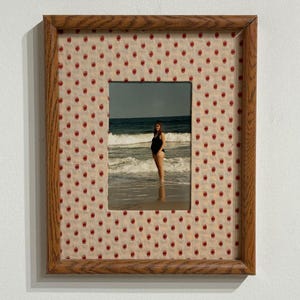 May include: A rectangular wooden picture frame featuring a light beige mat with a repeating pattern of small red handprints. The frame contains a photograph of a person in a black swimsuit standing in the ocean surf with waves behind them.