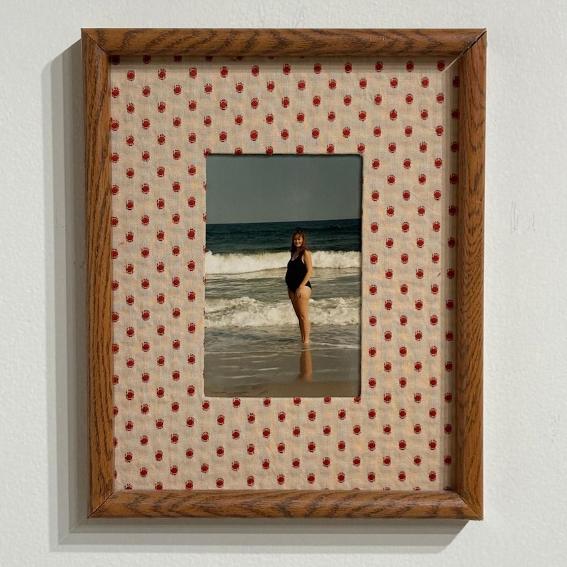 May include: A rectangular wooden picture frame featuring a light beige mat with a repeating pattern of small red handprints. The frame contains a photograph of a person in a black swimsuit standing in the ocean surf with waves behind them.
