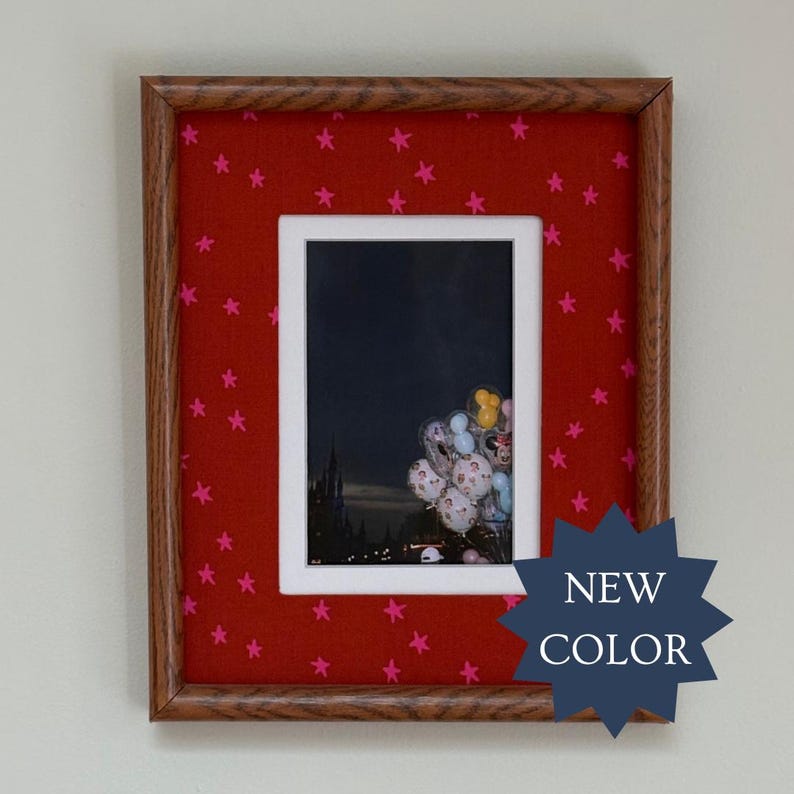 May include: A rectangular picture frame with a wood grain finish and a red mat featuring a pattern of small pink stars. The frame contains a photograph of a dark castle silhouette at night with a cluster of colorful balloons in the foreground.