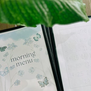 Op de afbeelding: Een wit en blauw ge&iuml;llustreerd menu met de tekst "morning menu" en een blauwe vlinder. Het menu is open naar een blanco pagina met het woord "juni" en gestippelde lijnen om de nummers 1 tot en met 30 te schrijven.
