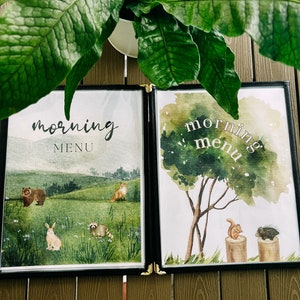Op de afbeelding: Twee ingelijste aquarel-illustraties met de tekst "morning menu" op elk. De linker illustratie toont een beer, een vos, een konijn en een wasbeer in een groene bosomgeving. De rechter illustratie toont een eekhoorn en een stekelvarken op boomstronken in een bosomgeving.