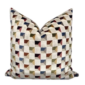May include: A square cushion with a geometric pattern. The cushion has a repeating design of three-dimensional squares in beige, brown, blue, green, and burgundy. The cushion is made of a soft, textured fabric. Measurements are not available.