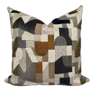 May include: A square throw cushion featuring a textured, multi-coloured geometric pattern. The design consists of various shapes in shades of cream, beige, dark brown, charcoal grey, and olive green, creating a complex, interlocking mosaic appearance.