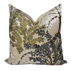 May include: A square throw pillow featuring an intricate embroidered botanical pattern of fern leaves in shades of olive green, dark charcoal, and metallic silver thread set against a light beige, textured fabric background.