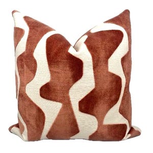 May include: A decorative throw pillow with a geometric pattern. The pillow features a rust-colored background with a white, abstract design. The pillow is square-shaped and made of a soft, textured fabric.