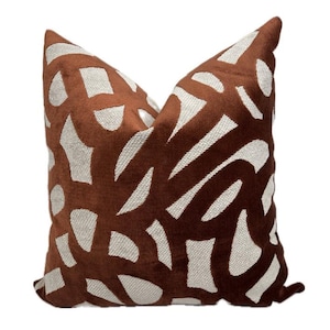 Puede incluir: Un cojín decorativo con un fondo marrón y un patrón repetitivo de formas blanquecinas. El cojín tiene un aspecto suave y texturizado y está diseñado para la decoración del hogar.