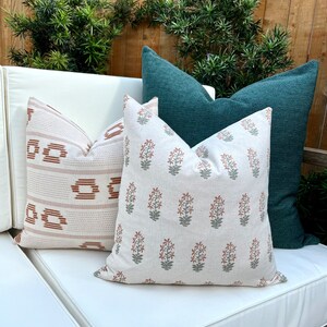 May include: Three throw pillows on a white couch. The pillows are a cream color with a rust and green floral pattern, a cream color with a rust and white geometric pattern, and a dark teal color.