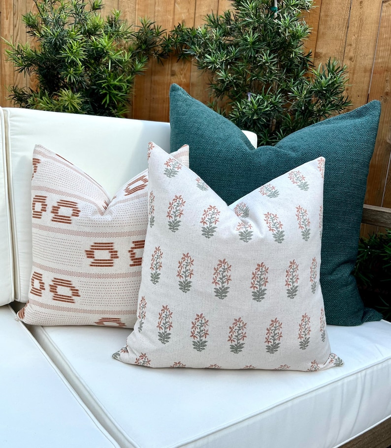 May include: Three throw pillows on a white couch. The pillows are a cream color with a rust and green floral pattern, a cream color with a rust and white geometric pattern, and a dark teal color.