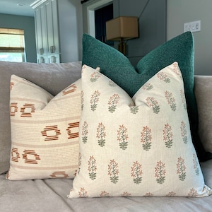 May include: Three throw pillows on a couch. The pillow in the front is white with a floral pattern in shades of green and orange. The middle pillow is white with a geometric pattern in shades of orange and brown. The back pillow is a dark green.
