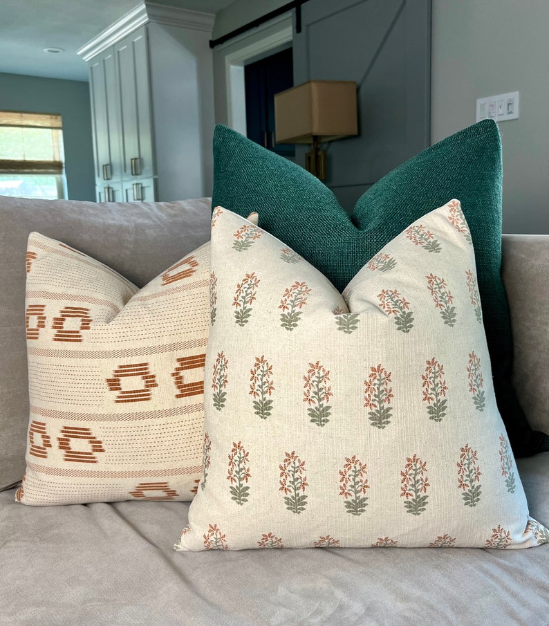May include: Three throw pillows on a couch. The pillow in the front is white with a floral pattern in shades of green and orange. The middle pillow is white with a geometric pattern in shades of orange and brown. The back pillow is a dark green.