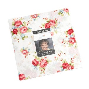 May include: A white fabric with a floral pattern featuring red roses and green leaves. The fabric has a subtle pink and blue background. The fabric is labeled "moda fabrics & supplies" and has a small photo of a woman in the center.