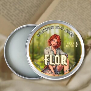 May include: A silver-colored tin of soy wax candle with the text "SPLINTERED BOND SERIES" and "FLOR". The label features an illustration of a woman with red hair. The text "SOUTHERN SPICED CIDER" and "MERRI BRIGHT" are also visible.
