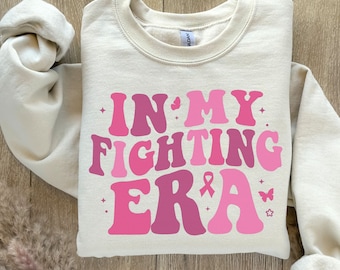 Peut inclure: Un sweat-shirt beige avec le texte "IN MY FIGHTING ERA" en lettres roses et violettes. Le texte comprend un ruban rose et des papillons.