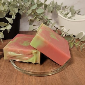 May include: Two bars of handmade soap, one green and pink swirl and one pink and green swirl, on a clear glass dish. The soap is on a wooden surface.