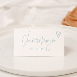 May include: White plates with a white card on top that says "Cheeseburger SLIDERS" with a blue heart next to the word "Cheeseburger".