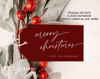 May include: A burgundy red gift tag with white text that reads "merry christmas" and "LOVE, THE JOHNSONS". The tag is hanging from a sprig of greenery with red berries.