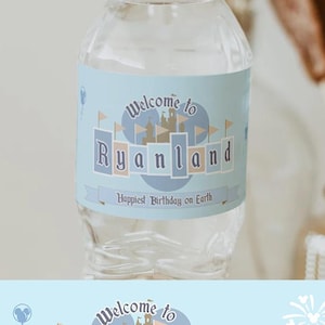 May include: A clear water bottle with a light blue label that reads "Welcome to Ryanland, Happiest Birthday on Earth." The label has a castle design. The bottle sits on a wooden base.