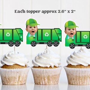 Op de afbeelding: Drie cupcakes met witte glazuur, elk versierd met een groene vuilniswagen-topper met een babygezicht. De toppers zijn ongeveer 6,6 cm x 5,1 cm groot. De cupcakes zitten in witte papieren vormpjes.
