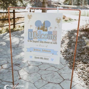 May include: A welcome sign for a baby shower, featuring a castle design and the text "Welcome to the Happiest Baby Shower on earth in honor of Jessica Johnson." The sign is displayed on a copper-colored metal stand. The date March 25th, 2018 is also included.