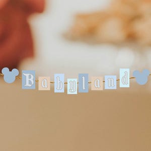 May include: A banner with the word "Babyland" spelled out in pastel-colored rectangular blocks. The letters are in a decorative font, and the banner is strung with a gold-colored string. Light blue Mickey Mouse head shapes are on each end.
