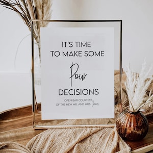 May include: A gold-framed sign with the text "It's time to make some Pour Decisions. Open bar courtesy of the new Mr. and Mrs. Jones." The sign is sitting on a wooden surface with a brown fabric and dried pampas grass.