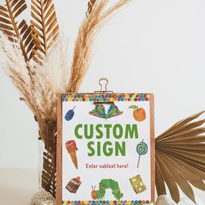 Peut inclure: Un presse-papiers en bois avec les mots "CUSTOM SIGN" en vert, avec des illustrations color&eacute;es d'aliments. Le panneau est maintenu par une pince dor&eacute;e et repose sur une base en bois. Des plantes s&eacute;ch&eacute;es et des &eacute;l&eacute;ments d&eacute;coratifs entourent le panneau.