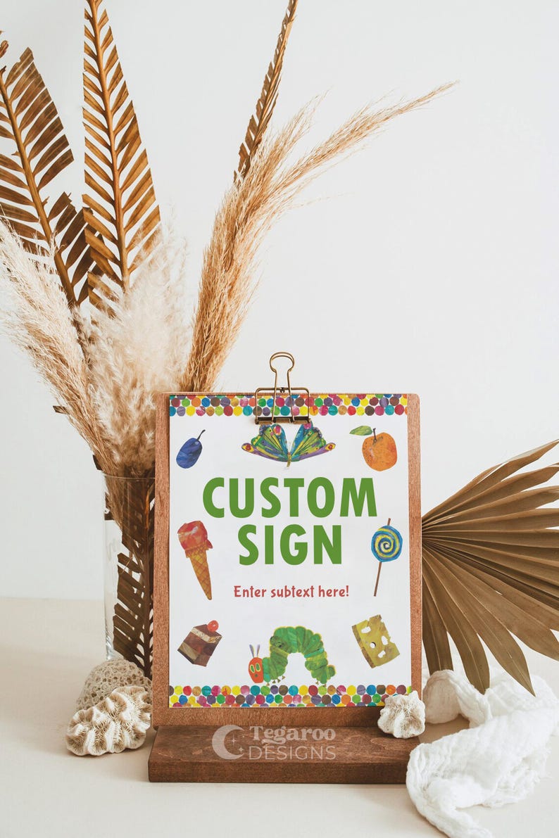 Peut inclure: Un presse-papiers en bois avec les mots "CUSTOM SIGN" en vert, avec des illustrations color&eacute;es d'aliments. Le panneau est maintenu par une pince dor&eacute;e et repose sur une base en bois. Des plantes s&eacute;ch&eacute;es et des &eacute;l&eacute;ments d&eacute;coratifs entourent le panneau.