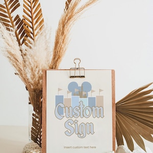 Może przedstawiać: Drukowana tabliczka z grafiką niebieskiego i brązowego zamku oraz tekstem "Custom Sign" z napisem "Insert custom text here" poniżej. Tabliczka stoi na drewnianym stojaku z napisem "Tegaroo Designs" poniżej.