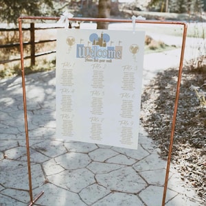 May include: A copper-colored metal frame holds a welcome sign with a Disney theme. The sign features the word "Welcome" and a list of names organized by table numbers. The sign is decorated with Mickey Mouse silhouettes and a castle.