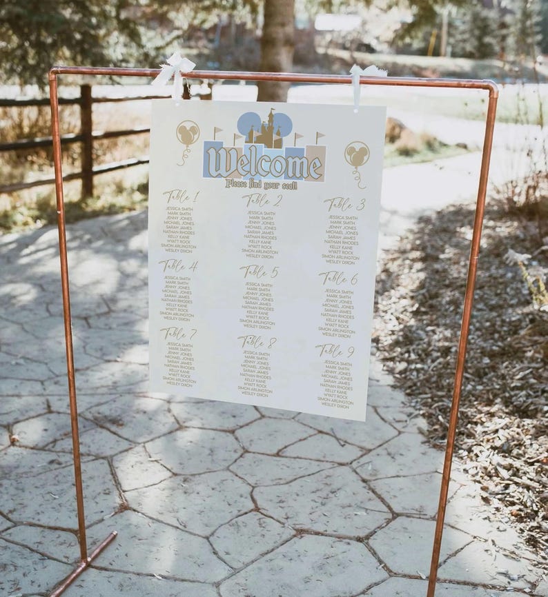 May include: A copper-colored metal frame holds a welcome sign with a Disney theme. The sign features the word "Welcome" and a list of names organized by table numbers. The sign is decorated with Mickey Mouse silhouettes and a castle.