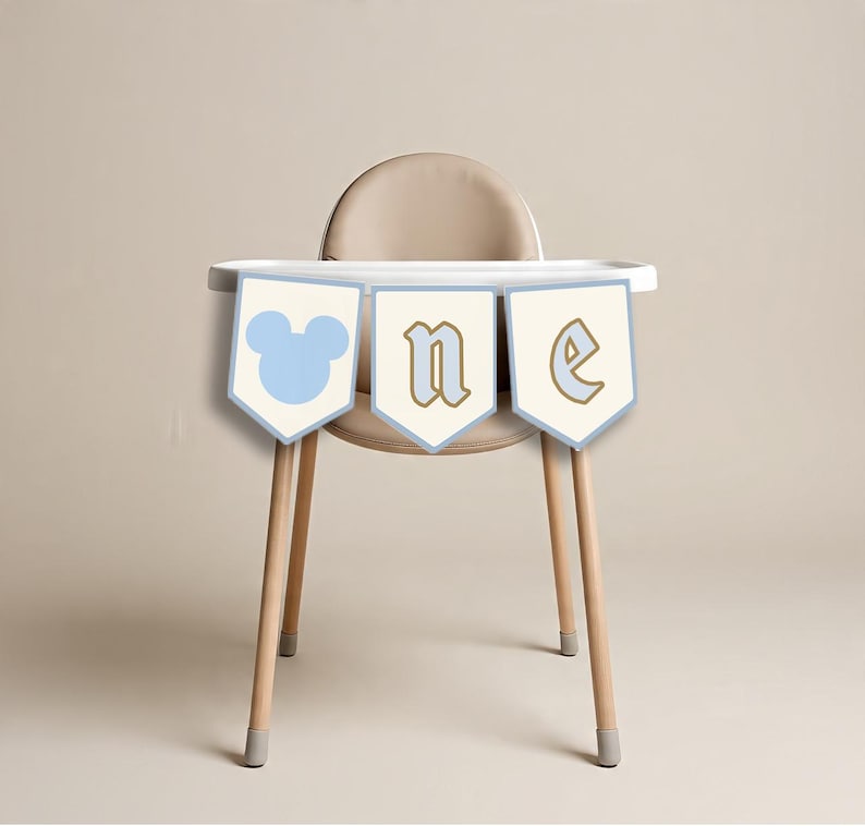 Puede incluir: Una pancarta con la palabra "one" y una forma de cabeza de Mickey Mouse, en azul claro y dorado, cuelga de una trona. La pancarta es de tela blanca con un borde azul claro. La trona tiene un asiento beige y patas de madera clara.