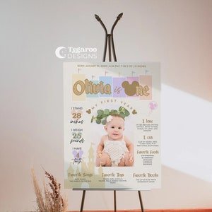 Op de afbeelding: Een pastelkleurig eerste verjaardagsbord met een foto van een babymeisje. Het bord bevat de naam van de baby, Olivia, en details over haar eerste jaar, inclusief haar lengte, gewicht en favoriete dingen. Het bord is versierd met een gouden glitter-&eacute;&eacute;n, een Mickey Mouse-hoofd en een bloemenkrans.