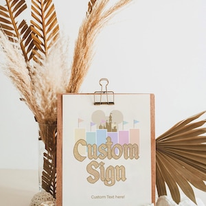 May include: A wooden sign with a gold glitter castle graphic and the text "Custom Sign" with the words "Custom Text here!" below. The sign is on a wooden stand with the text "Tegaroo Designs" on the front.