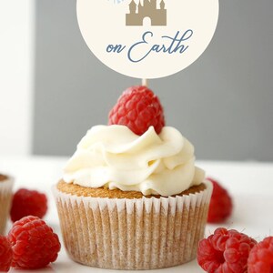 May include: A cupcake with white frosting and a fresh raspberry on top. A decorative sign with the text "Happiest Baby shower on Earth" and a castle illustration is placed on top of the cupcake. Several raspberries are scattered around the cupcake.