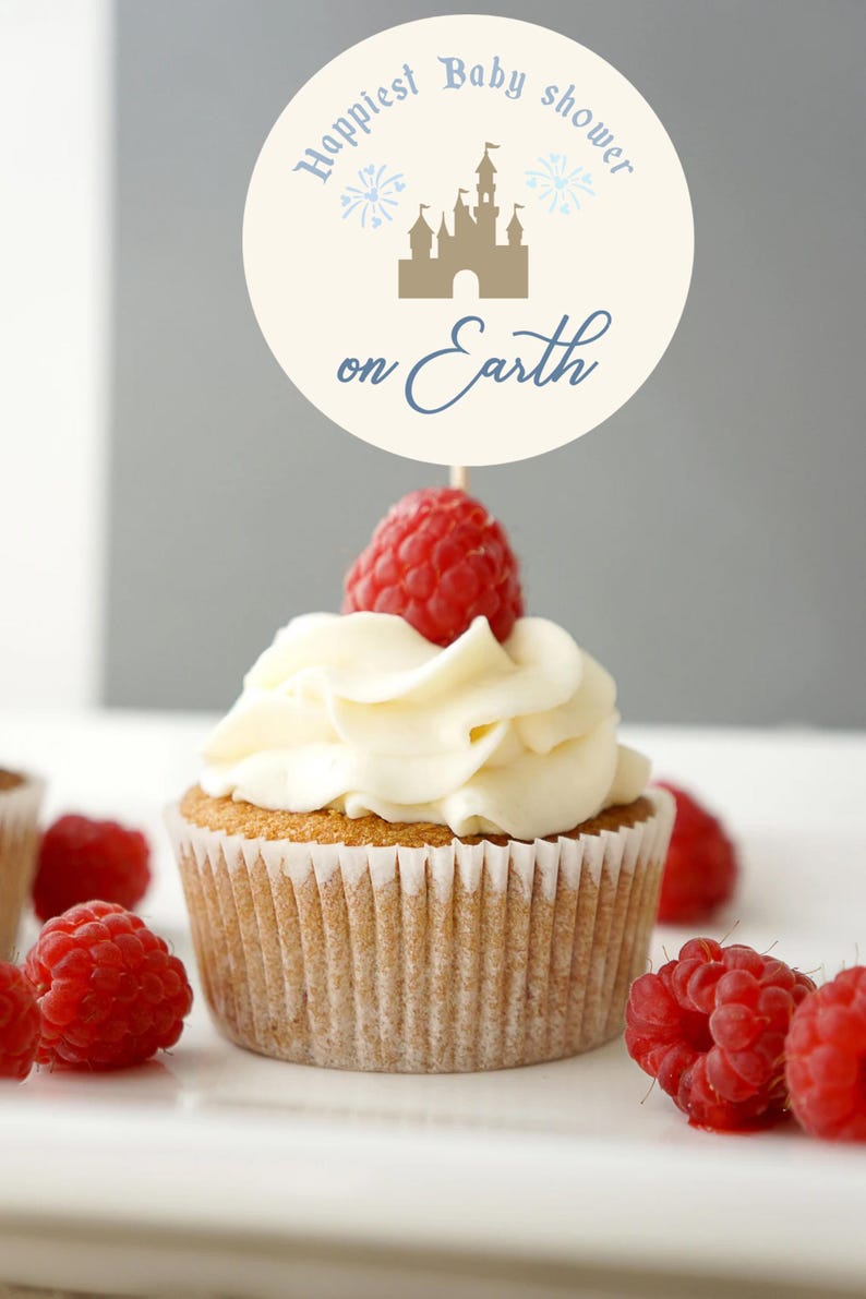 May include: A cupcake with white frosting and a fresh raspberry on top. A decorative sign with the text "Happiest Baby shower on Earth" and a castle illustration is placed on top of the cupcake. Several raspberries are scattered around the cupcake.
