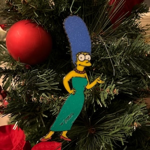 May include: A Christmas tree decoration of Marge Simpson from The Simpsons. The ornament shows Marge in her green dress and tall blue hair. The decoration is hanging on a Christmas tree with a red bauble and red poinsettia.