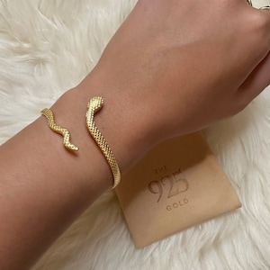 May include: A gold snake-shaped bracelet with a textured finish. The bracelet is on a person's wrist and is accompanied by a small box with the text "THE 925 GOLD" on it.