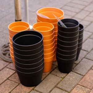 May include: A stack of orange plastic plant pots and a stack of black plastic plant pots on a brick surface.