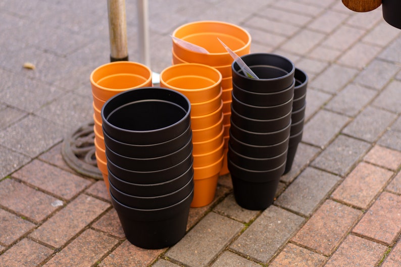 May include: A stack of orange plastic plant pots and a stack of black plastic plant pots on a brick surface.
